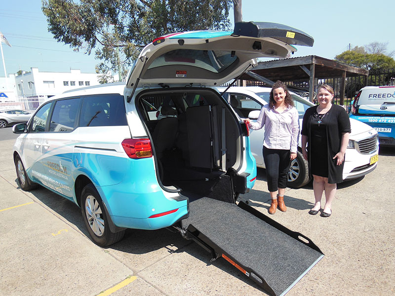 Our friendly NSW Sales managers Shauny Van Mullekom & Carolyn Hurst standing next to an open rear door of an accessibility van with a lowered wheelchair ramp.
